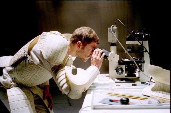 Image from Il Combattimento - A man in padded armor looks through a microscope intently
