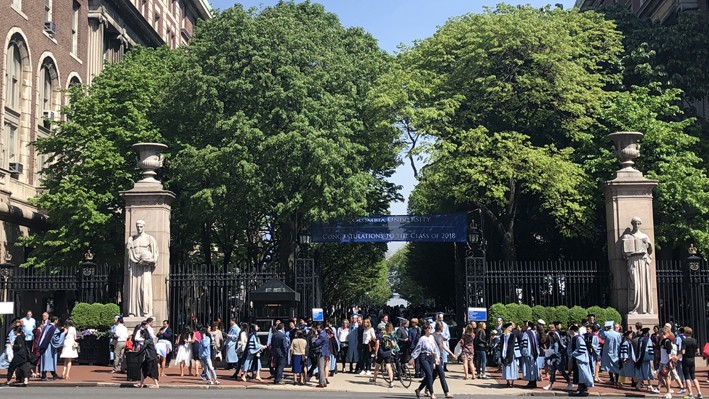 Graduates in light blue gowns, along with friends and family, take photos in front of the main Columbia University gates